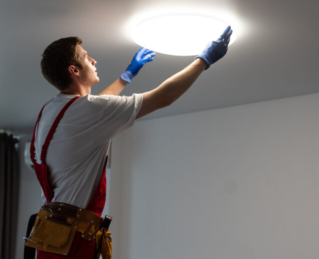 A person in work attire and gloves adjusts a ceiling light fixture in a room with white walls and a curtain.
