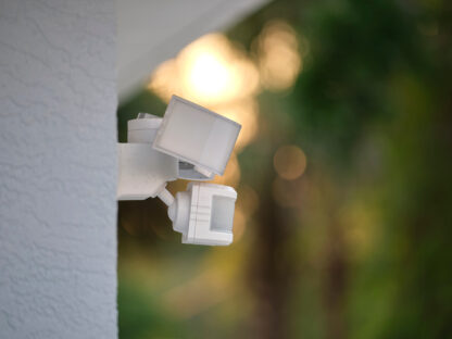 A white motion sensor light mounted on a white textured wall, with a blurred background of greenery and soft sunlight.