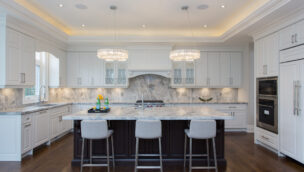 A spacious modern kitchen with a large marble island, three gray barstools, white cabinetry, and two crystal chandeliers.