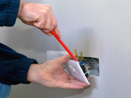 Person using a red screwdriver to install or repair a wall electrical outlet, with exposed wires visible inside the wall cutout.