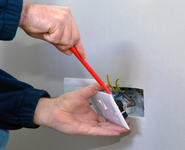 Person using a red screwdriver to install or repair a wall electrical outlet, with exposed wires visible inside the wall cutout.