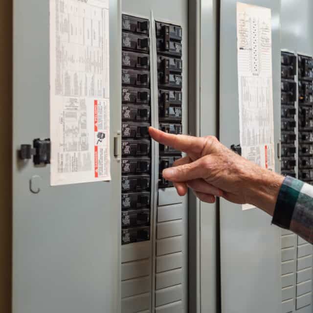 A person adjusts a breaker switch inside an open electrical panel mounted on a wall.