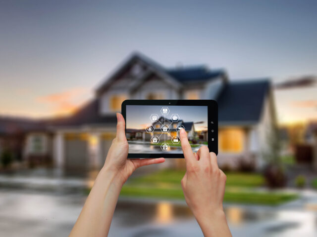 Person using a tablet to control smart home features, with a modern house in the background.