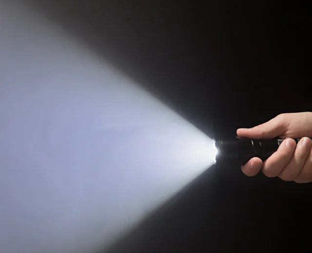 Black flashlight with a beam of light in male's hand isolated from right side of the frame on black background.