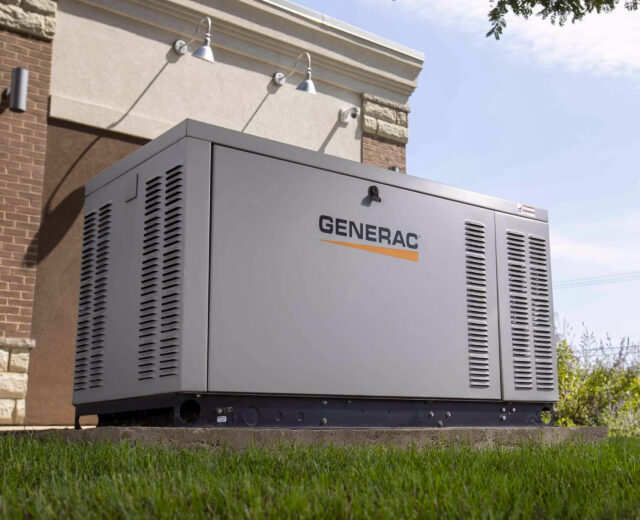Large Generac generator on a concrete pad beside a building, surrounded by grass, with trees partially visible above.