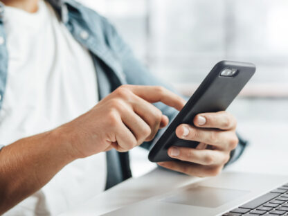 Person using a smartphone while seated at a desk with a laptop.