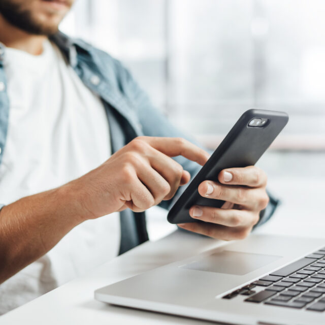 Person using a smartphone while seated at a desk with a laptop.