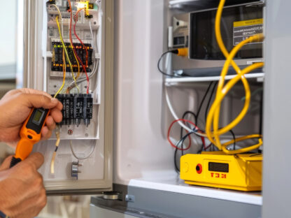 A person uses a handheld device to test wires in an electrical panel box, with various tools and a yellow voltmeter displayed.