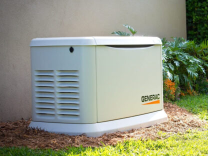 A Generac standby generator is installed next to a house, surrounded by mulch, with green grass and plants nearby.