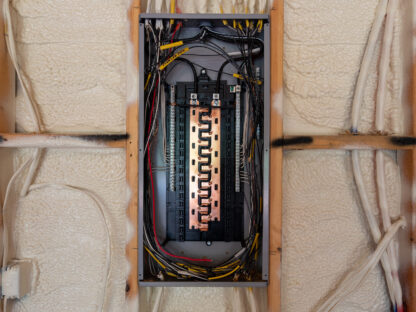 An open electrical panel with visible wiring and circuit breakers is set against a wall with insulation foam.